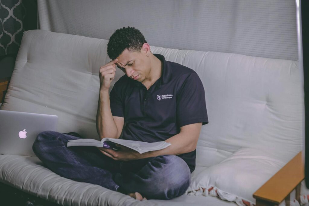 A student is relaxing on a sofa, studying with a book, and using a laptop indoors.