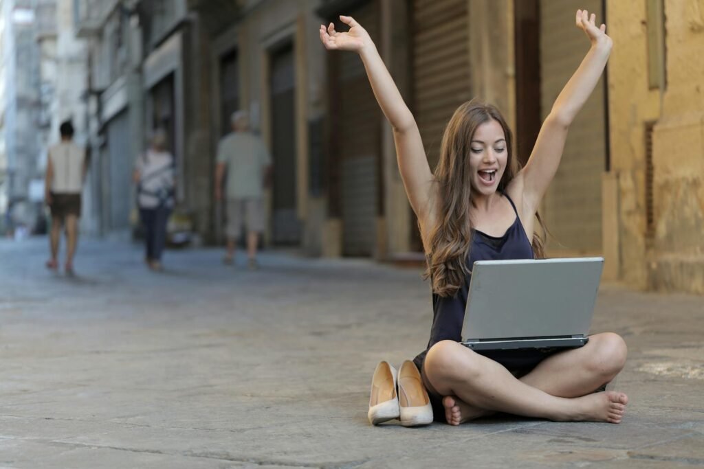 A cheerful woman sitting outdoors, celebrating success with arms raised while using a laptop.