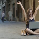 A cheerful woman sitting outdoors, celebrating success with arms raised while using a laptop.