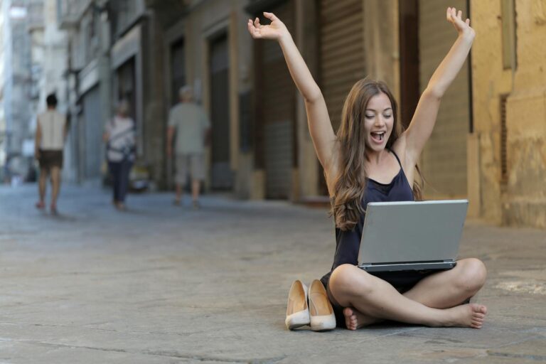 A cheerful woman sitting outdoors, celebrating success with arms raised while using a laptop.