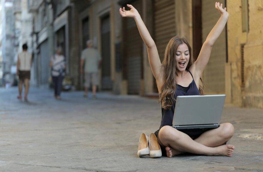 A cheerful woman sitting outdoors, celebrating success with arms raised while using a laptop.