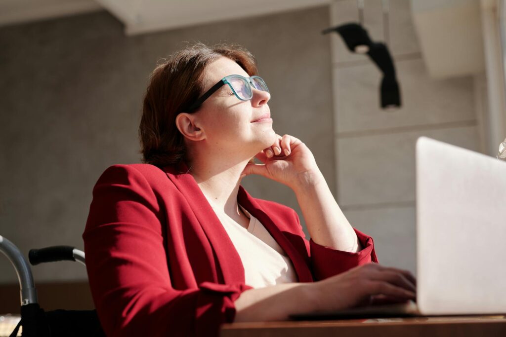 A woman in a wheelchair wearing a red suit and glasses, working on a laptop in a bright café.