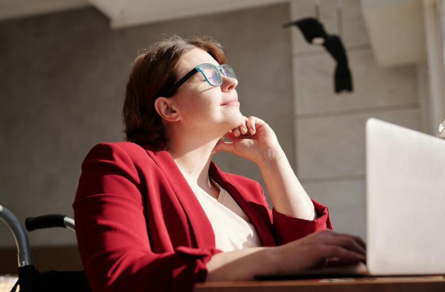 A woman in a wheelchair wearing a red suit and glasses, working on a laptop in a bright café.