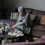 A Muslim woman in traditional attire is working on her laptop in a cozy living room, studying self-improvement.