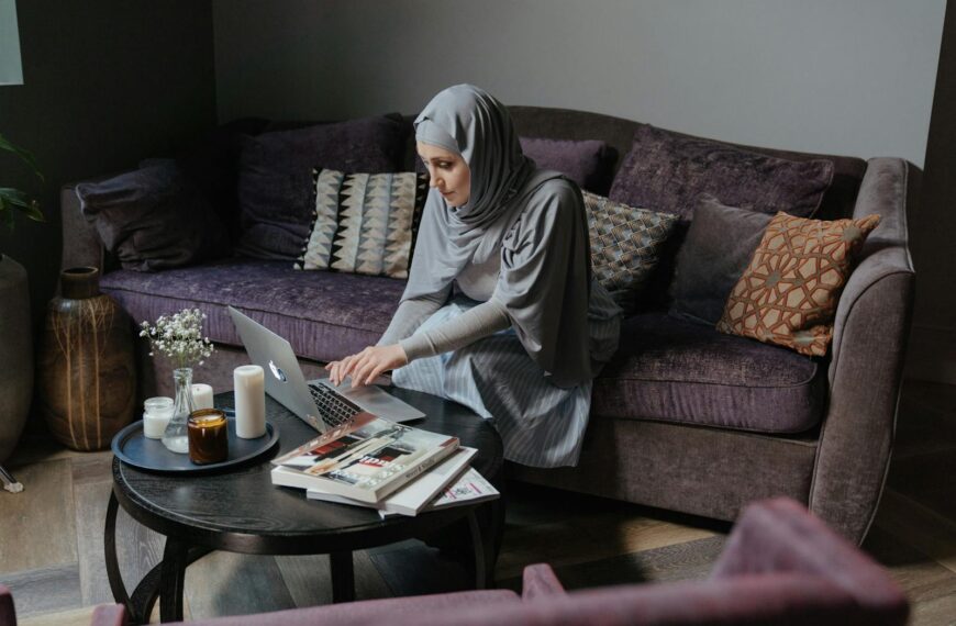 A Muslim woman in traditional attire is working on her laptop in a cozy living room, studying self-improvement.