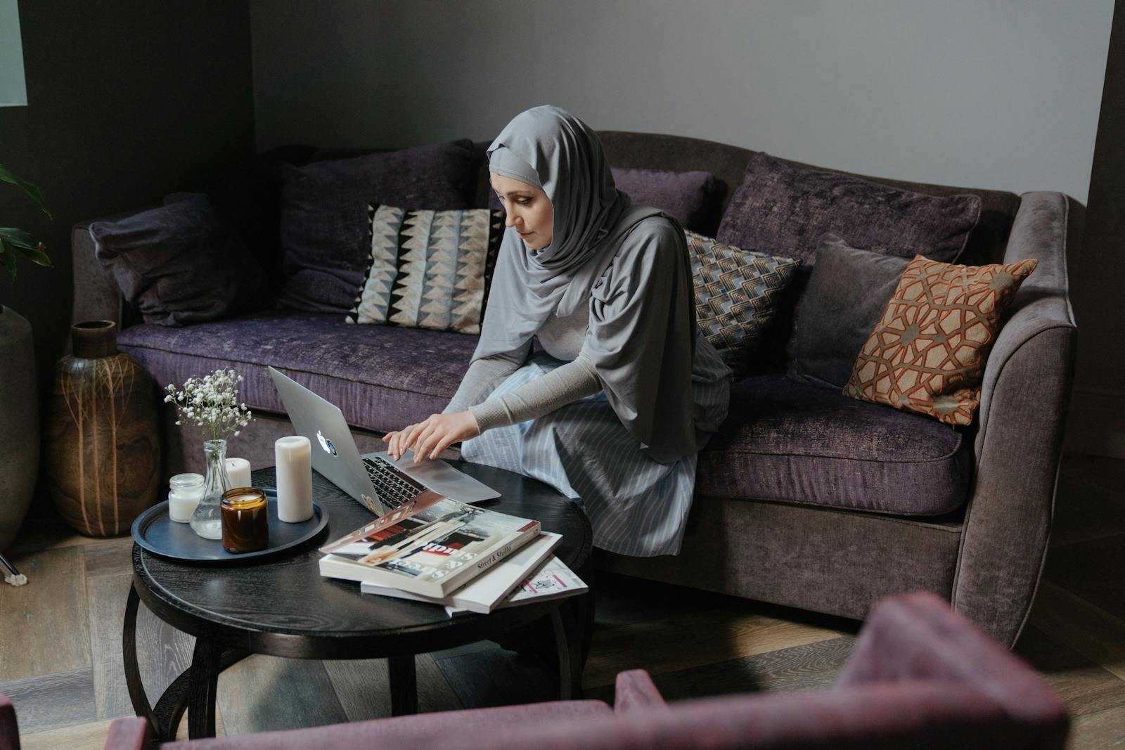 A Muslim woman in traditional attire is working on her laptop in a cozy living room, studying self-improvement.