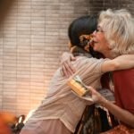 The older woman and younger woman share a heartfelt hug indoors, holding a gift.