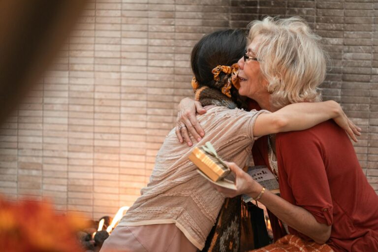 The older woman and younger woman share a heartfelt hug indoors, holding a gift.