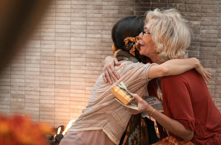 The older woman and younger woman share a heartfelt hug indoors, holding a gift.