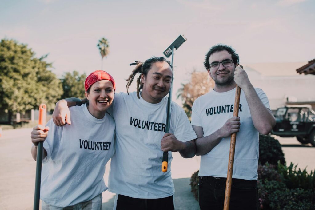 Three diverse volunteers are smiling and holding cleaning tools during a community service event.