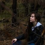 A woman in a black jacket sitting in the woods pondering a mindset shift