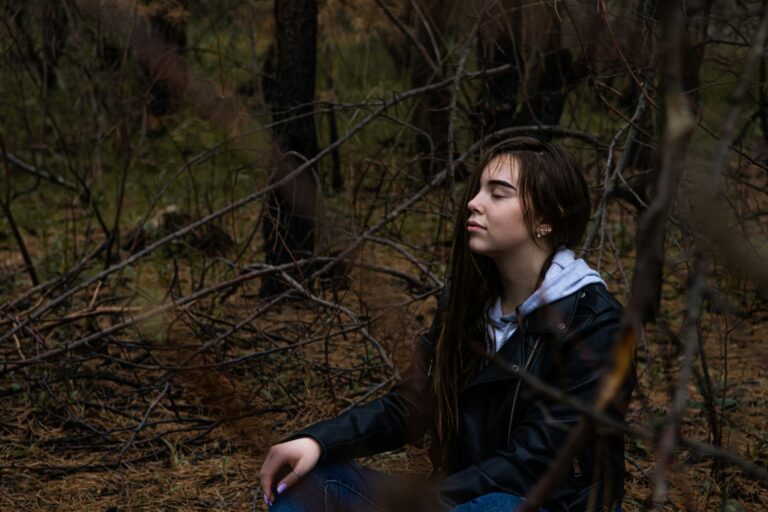 A woman in a black jacket sitting in the woods pondering a mindset shift